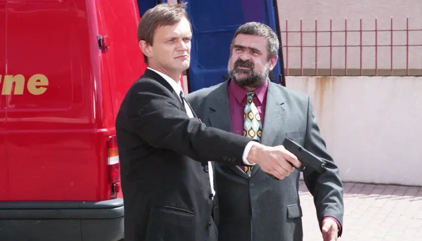 Gangster z filmu Kiler, w garniturze, celuje z pistoletu. Obok stoi mężczyzna w czarnym garniturze.