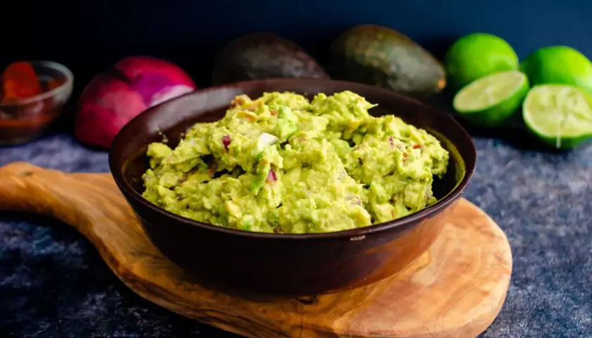 Miseczka guacamole z kawałkami cebuli i pomidorów, na drewnianej desce, w tle awokado i limonki.