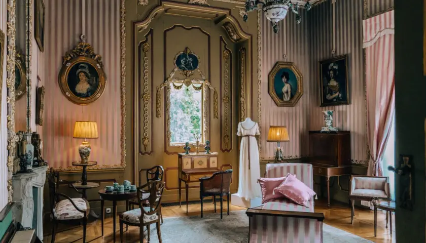 Eleganckie wnętrze pałacu w Nieborowie. W pokoju z różowo-białymi pasami na ścianach, zabytkowe meble, obrazy i manekin w sukni.