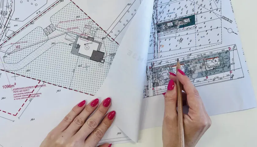 Rysunek techniczny z naniesionym projektem przyłącza wodociągowego. Dłoń z paznokciami malowanymi na różowo trzyma ołówek nad dokumentem.