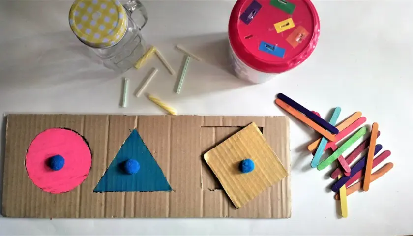Kolorowe zabawki DIY dla niemowlaka: sorter kształtów z kartonu, patyczki i słoik.