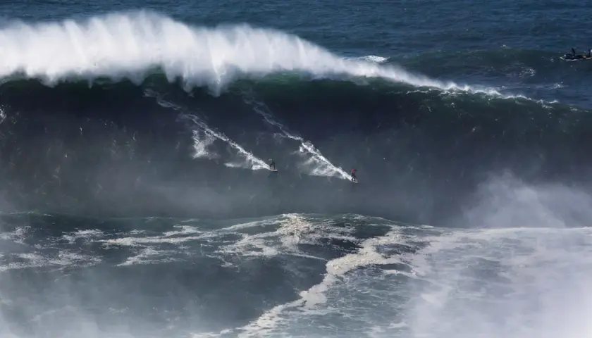 Surferzy ścigają się po gigantycznych nazare falach, tworząc białe smugi na ciemnym oceanie.