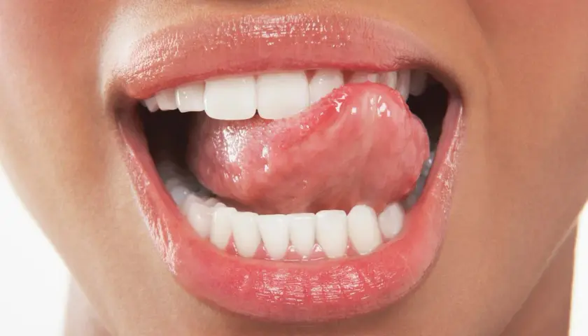 Labbra lucide e lingua che accarezza i denti ruvidi, un gesto sensuale che invita a scoprire.