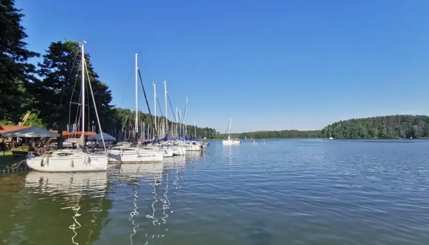 Żaglówki cumują w porcie w Rucianem Nidzie. Słoneczny dzień nad jeziorem, zalesiony brzeg w oddali.