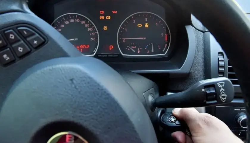 Ręka próbuje odpalić auto, ale kontrolki świecą. Kluczyk w stacyjce, a na desce rozdzielczej widoczne m.in. kontrolka silnika i ABS.