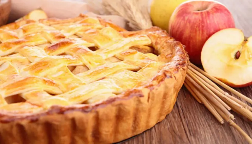 Tarta de manzana dorada con rejilla de masa, junto a manzanas frescas y palitos de canela.