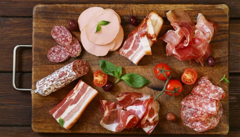 Assortiment de différents types de charcuterie : saucisson, jambon cru, bacon, mortadelle, accompagnés de tomates cerises et d'olives.