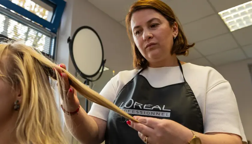 Fryzjerka w fartuchu L'Oréal Professionnel wykonuje podcinanie końcówek blond włosów.