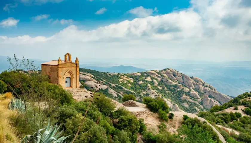 Pequeña ermita en la cima de una colina, con vistas a las impresionantes rutas de Montserrat y un paisaje montañoso bajo un cielo azul con nubes.