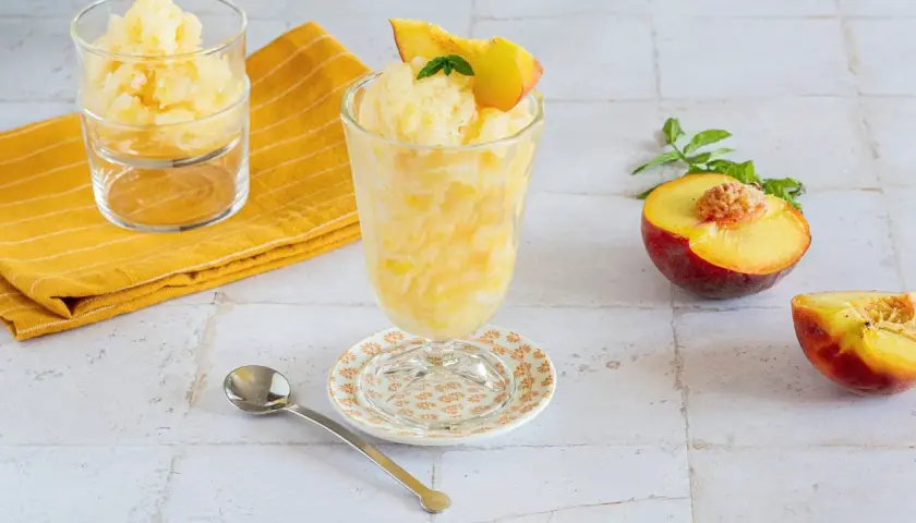 Rinfrescante granita alla pesca, guarnita con una fetta di pesca e menta. Accanto, pesche fresche e un cucchiaino.