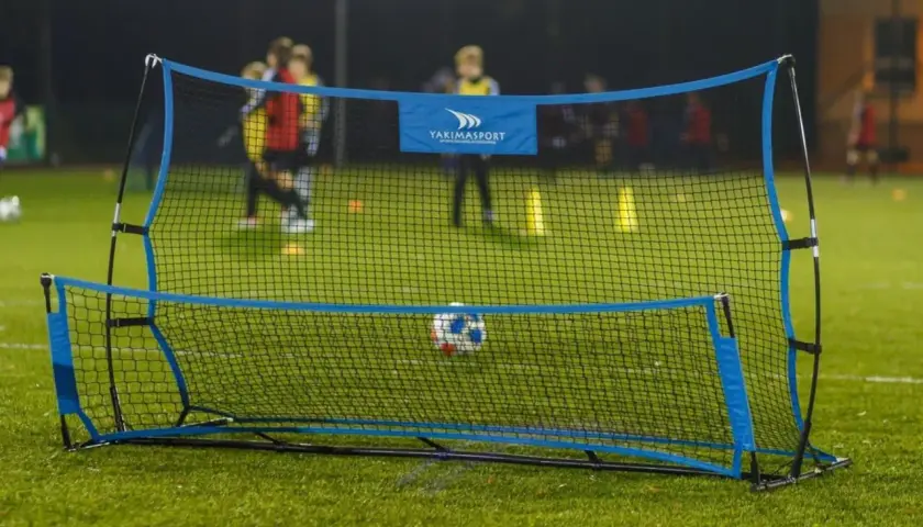 Piłkarski rebounder Yakimasport na zielonej murawie, z piłką w siatce. W tle trening młodych piłkarzy.