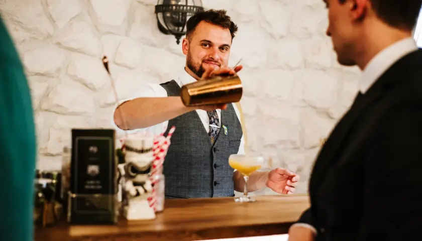 Uśmiechnięty barman w kamizelce nalewa drinka do kieliszka.