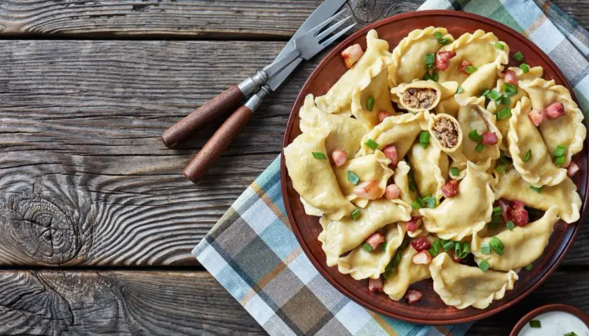 Pyszne pierogi z mięsem, posypane skwarkami i szczypiorkiem, czekają na talerzu. Obok sztućce i miseczka śmietany.