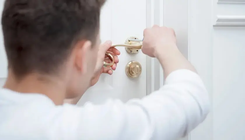 Un homme répare une poignée de porte dorée avec un tournevis sur une porte blanche.