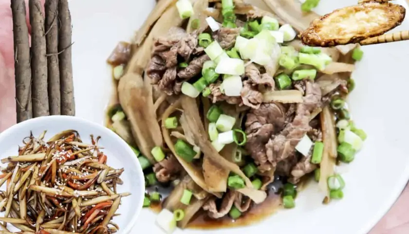 A delicious dish of braised burdock with thinly sliced beef and green onions, served on a white plate. A small bowl of burdock stir-fry is on the side.
