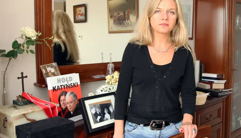 Kobieta z blond włosami w czarnej bluzce i jeansach, obok książka "Hołd Katyński" i ramka ze zdjęciem.