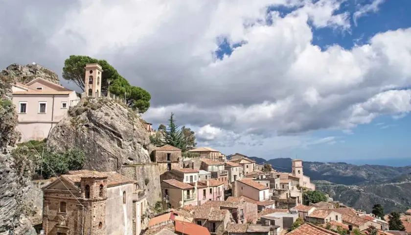 Charming towns in Calabria, Italy, cling to a rocky hillside under a dramatic sky. A bell tower stands sentinel over terracotta roofs.