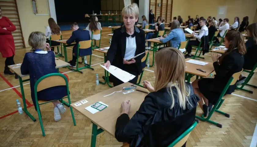 Napięcie w sali gimnastycznej, kiedy rozdawane są arkusze. Uczniowie czekają na wyniki egzaminu ósmoklasisty, a nauczycielka czuwa nad przebiegiem.