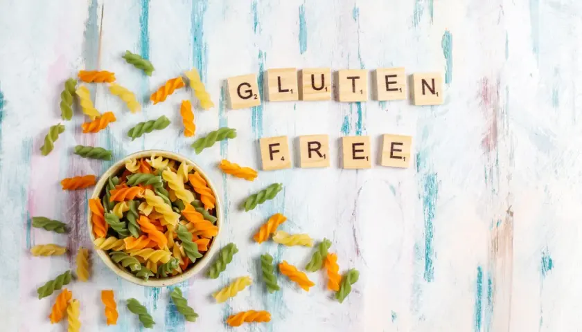 Kolorowy makaron w miseczce i rozsypane po stole, litery układające się w napis "GLUTEN FREE".