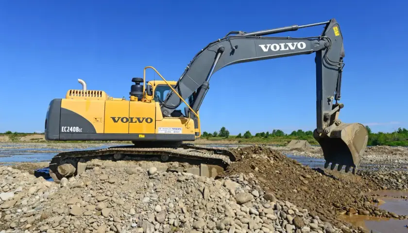 Żółta koparka Volvo EC240BLc pracuje na kamienistym brzegu rzeki. Zastanawiasz się, ile bierze koparka za godzinę?