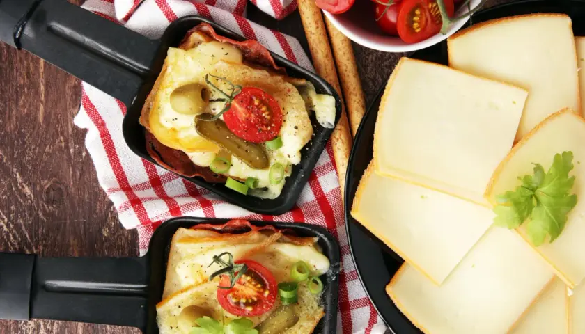 Deux poêlons de raclette fondue, garnie de tomates, cornichons et olives, accompagnés de fines tranches de fromage. Une variante gourmande !