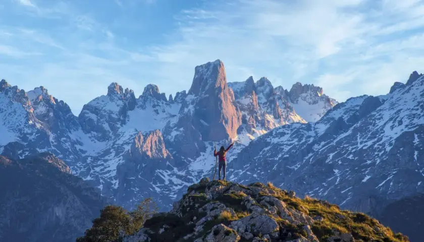 Persona en la cima de una montaña rocosa, con los brazos extendidos, contemplando los impresionantes picos nevados de los Picos de Europa. Un paisaje espectacular para descubrir qué ver.