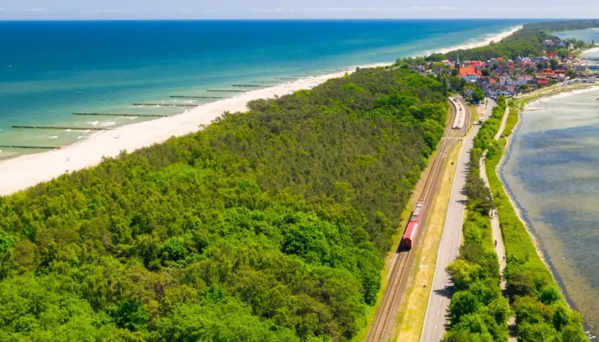 Czerwony pociąg jedzie wzdłuż wybrzeża. W tle widać plażę, morze i nadmorską miejscowość. Idealna podróż pociągiem nad morze.