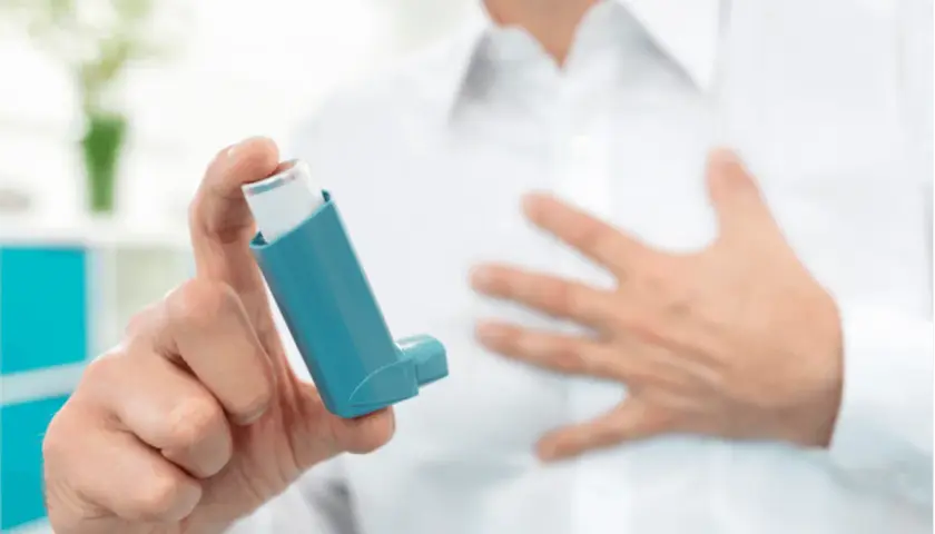 Mann hält Inhalator mit ipratropiumbromid. Seine andere Hand liegt auf der Brust, was auf Atemnot hindeutet.