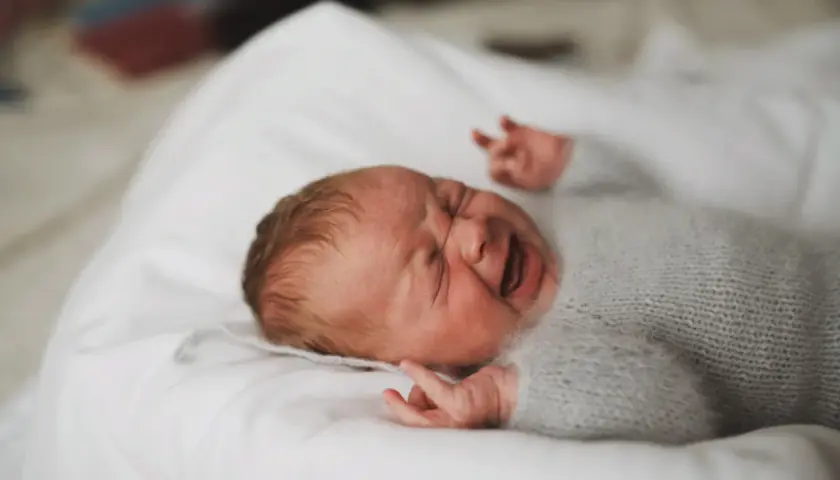En 1 månads bebis sover inte, med ögonen stängda och munnen öppen i ett skrik. Bebisen är insvept i en mjuk, grå stickad filt.
