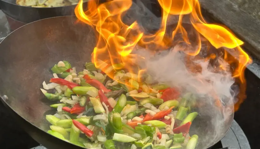Ogień płonie nad warzywami w woku, tworząc aromatyczny stir fry. W tle widać smażone ziemniaki.