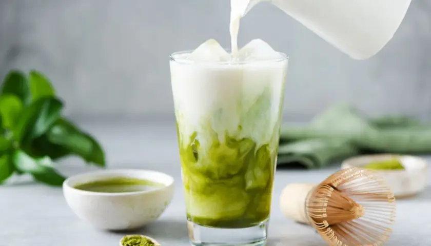 Eisiger Matcha Latte wird mit Milch übergossen. Die Zutaten für den Matcha Latte (kcal) sind auf dem Tisch verteilt.