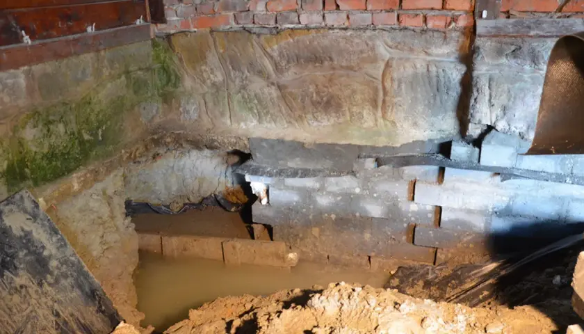 Prace nad hydroizolacją piwnicy od wewnątrz. Widoczny wykop, wilgotne ściany z cegły i kamienia, woda gruntowa.