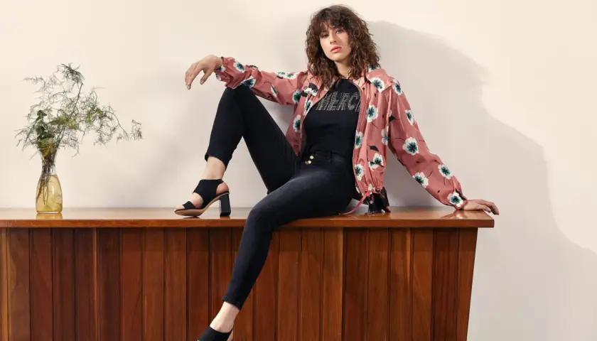 Mujer con rizos sentada en mueble de madera, luciendo distintos tipos de estilos de vestir: chaqueta floral, camiseta con brillos y jeans negros.