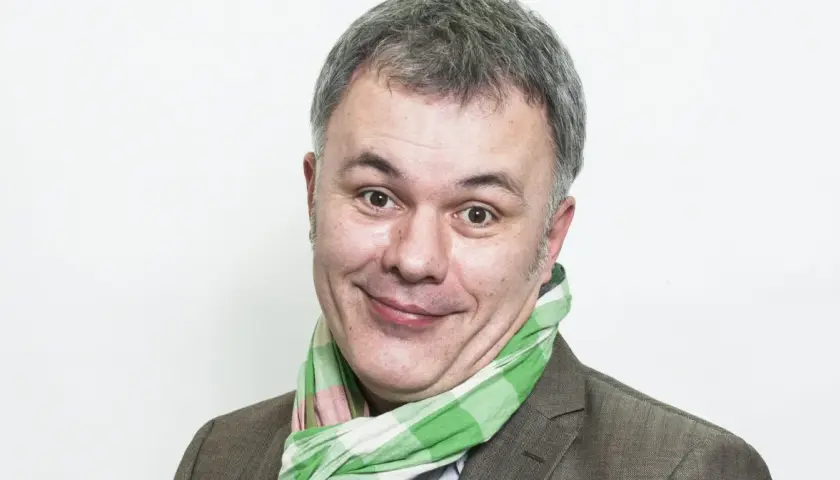 Uśmiechnięty mężczyzna w kratkowanym szaliku, znany jako Mazurek z RMF FM, z szarymi włosami i brązową marynarką.