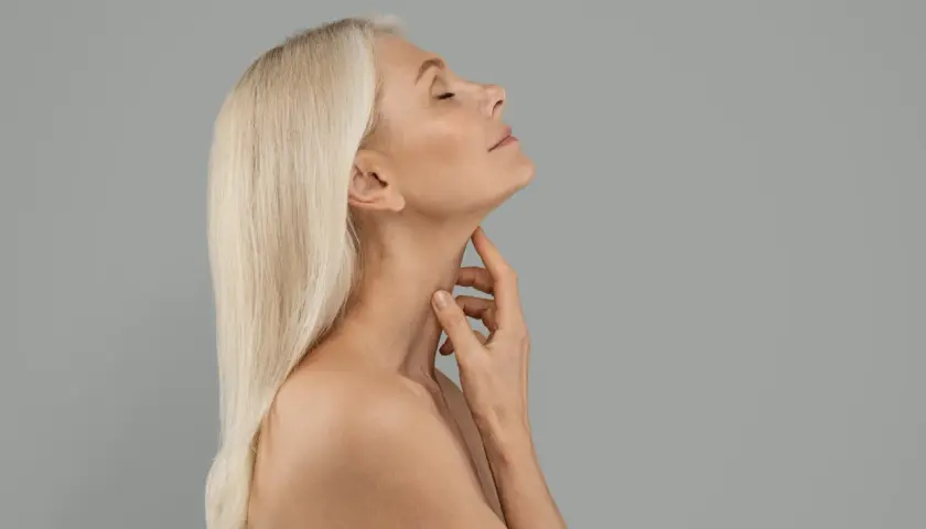 Äldre kvinna med blont hår rör vid sin hals, en bild som kan associeras med en kalkonhals operation.