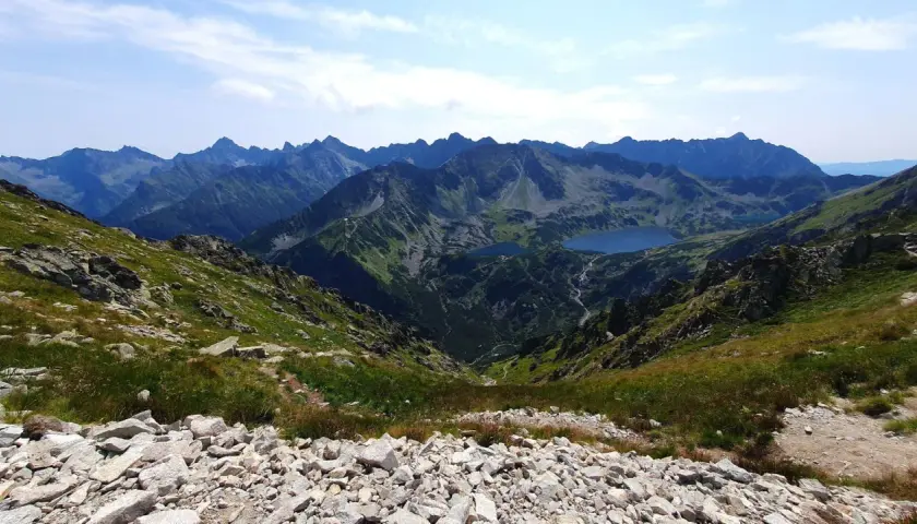 Widok na Tatry z kamienistego zbocza, z jeziorami w dolinie. Szlak turystyczny prowadzi przez malownicze **krzyżne** krajobrazy.