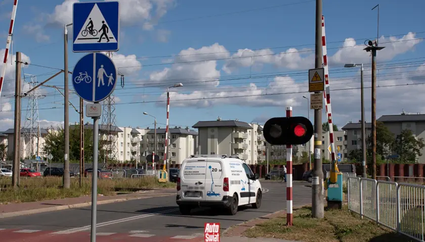 Czerwone światła na przejeździe kolejowym sygnalizują zatrzymanie. Znak informuje o przejściu dla pieszych i rowerzystów.