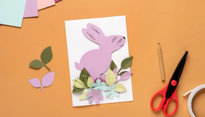 Uroczy różowy króliczek z papieru na kartce wielkanocnej DIY, otoczony kolorowymi kwiatami i liśćmi. Obok nożyczki, ołówek i taśma.