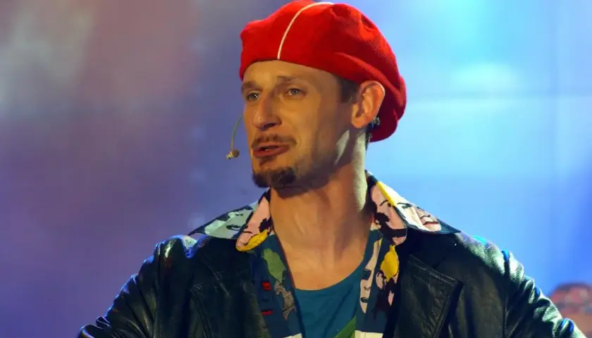 Michał Wójcik w czerwonej beretce i kolorowej koszuli, z mikrofonem przy ustach, na tle niebiesko-fioletowego światła.