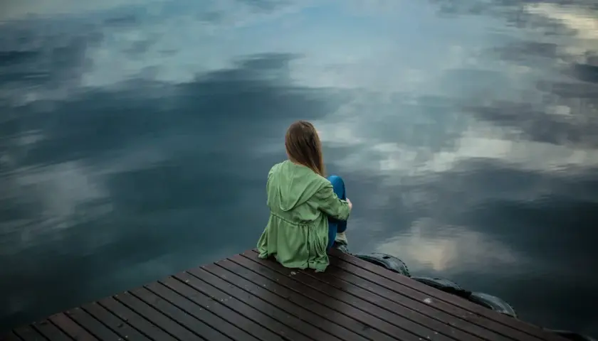 Ein Mädchen sitzt allein auf einem Holzsteg am Wasser. Sie wirkt nachdenklich, als ob sie nicht alleine sein kann.