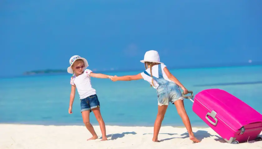 Due bambine si tengono per mano su una spiaggia tropicale, una tira una valigia rosa. Inizia la loro vacanza famiglia mare.