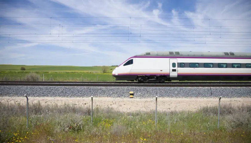 Un moderno tren blanco con detalles morados avanza por las vías. El paisaje verde y el cielo azul sugieren un viaje rápido de A Coruña a Madrid.