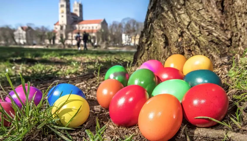 Bunte Ostereier liegen im Gras vor einem Baum, im Hintergrund eine Kirche. So feiert man an Ostern die Auferstehung.