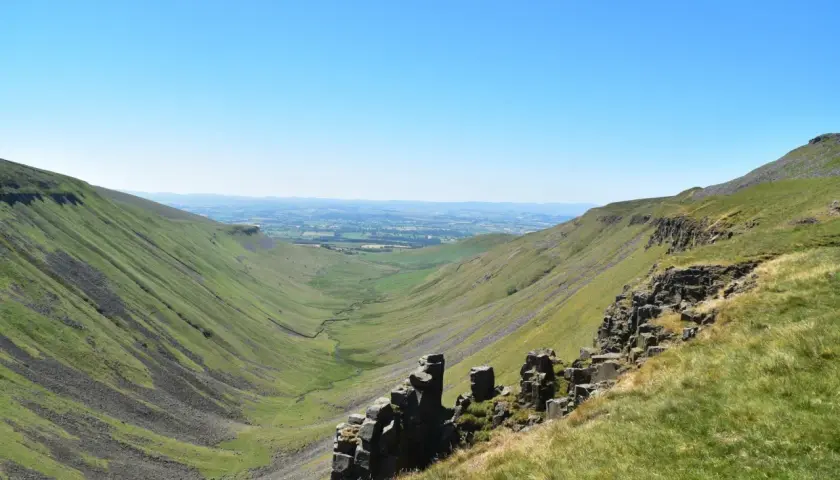 A vast green valley carved by a winding stream, with rocky outcrops in the foreground. This is a classic view along the Pennine Way, under a clear blue sky.