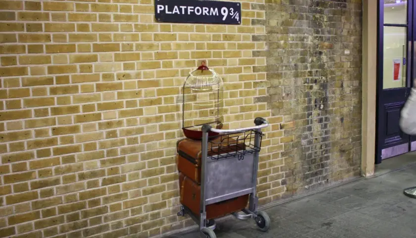 Wózek z bagażami i klatką dla ptaków przy ścianie z napisem "Platform 9 3/4" - magiczne miejsce jak z Kings Cross Harry Potter.