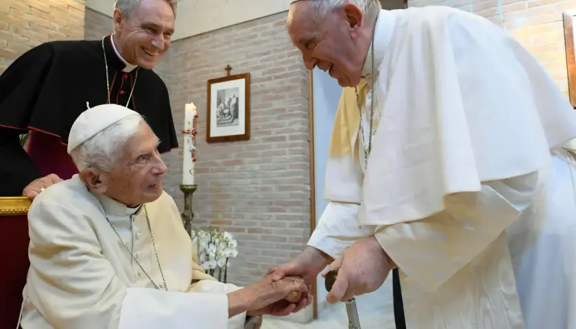 Papst Franziskus und ein Bischof besuchen Papst Benedikt. Sie reichen sich die Hände und lächeln.