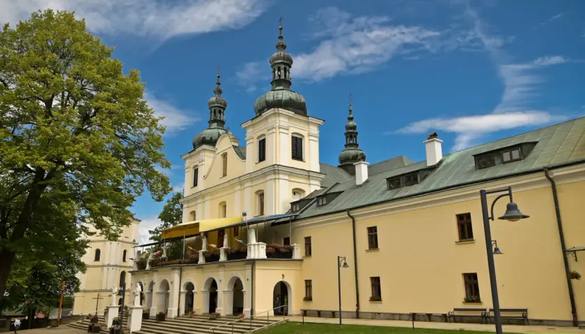 Jasny budynek sanktuarium z kopułami i krzyżami na wieżach, otoczony zielenią. Kalwaria Pacławska dróżki zapraszają do modlitwy.