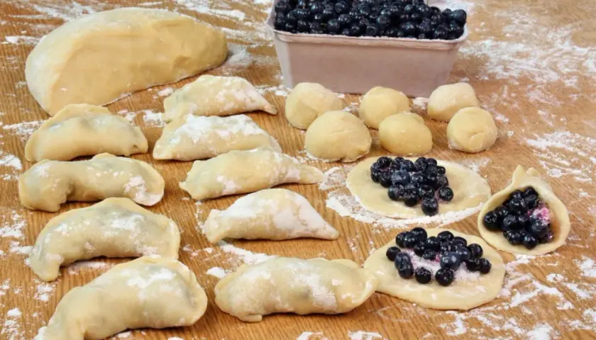 Przygotowania do tego, jak zrobić pierogi z jagodami: ciasto, kulki ciasta, gotowe pierogi i te w trakcie lepienia z jagodami.