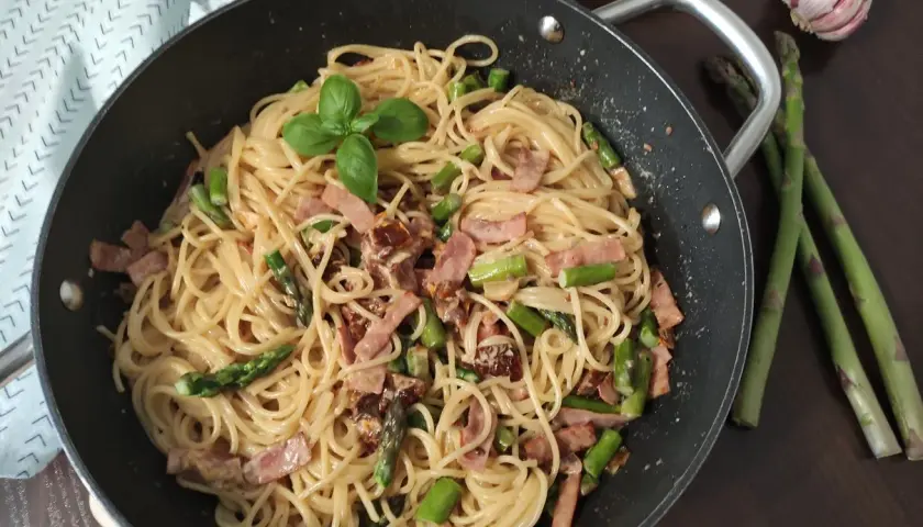 Pyszne spaghetti ze szparagami i boczkiem, udekorowane listkiem bazylii, czeka w patelni. Obok leżą świeże szparagi i czosnek.