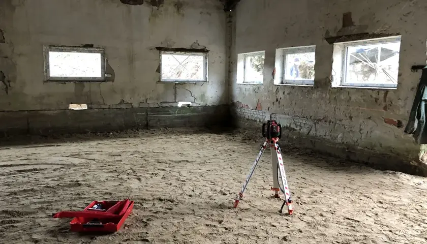 Na piaszczystym podłożu stoi niwelator geodezyjny, gotowy do pracy. W tle widać okna i ściany, a na ziemi leży czerwona skrzynka. To przygotowania do wylania posadzki z **gruntu do anhydrytu**.